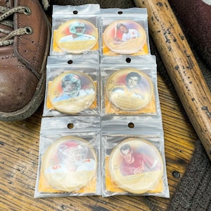 Peut inclure: Six pièces de collection sportives circulaires présentant des portraits d'athlètes dont Dan Marino, Hank Aaron, Tom Brady, Bobby Orr, Patrick Mahomes et Gordie Howe, chacune scellée individuellement dans un sachet plastique avec un carton jaune.