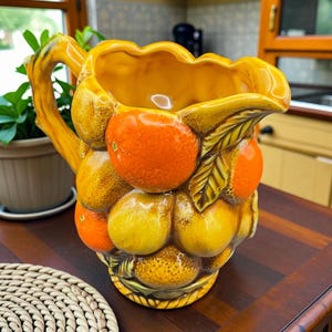 Peut inclure: Pichet en céramique avec un motif de fruits. Le pichet est jaune avec des accents de fruits orange et jaunes. La poignée a la forme d'une branche. Le pichet est posé sur une surface en bois foncé.