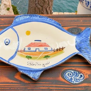 Peut inclure: Un plat en céramique en forme de poisson avec un intérieur blanc et une bordure bleue. Le plat présente une scène peinte d'une maison avec un toit rouge, un soleil jaune et le mot "Portugal".