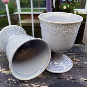 May include: Two pale blue ceramic goblets with a floral design on a distressed wooden surface. One goblet is upright, the other is tilted. The goblets have a pedestal base and a wide rim.