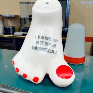 May include: A white ceramic foot-shaped souvenir figurine with red painted toenails. The text printed on the top of the foot reads, I WALKED MY FEET OFF IN WILDWOOD, N.J. The object has a smooth, glossy finish and a small hole at the top.