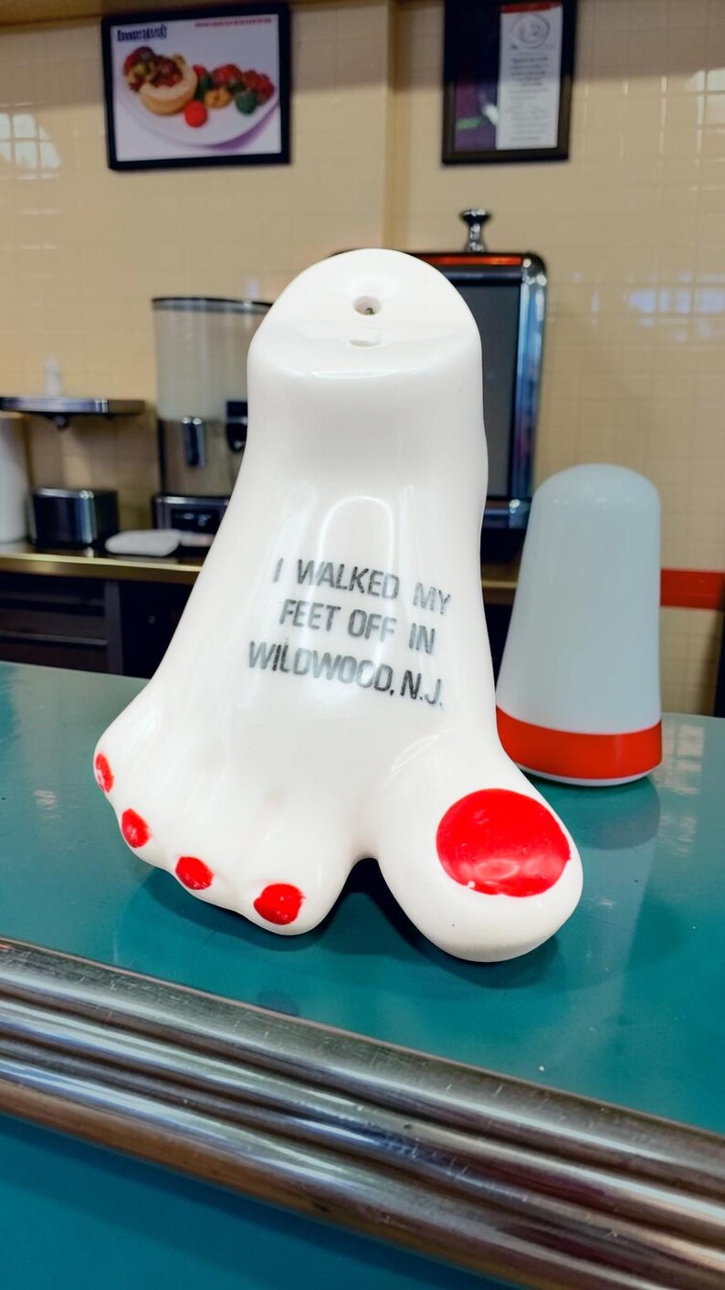 May include: A white ceramic foot-shaped souvenir figurine with red painted toenails. The text printed on the top of the foot reads, I WALKED MY FEET OFF IN WILDWOOD, N.J. The object has a smooth, glossy finish and a small hole at the top.