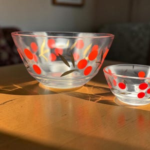 Peut inclure: Deux bols en verre transparent de tailles différentes, chacun présentant un motif répétitif de formes ovales rouge vif et de fines touches vert foncé en forme de feuilles peintes sur la surface extérieure des récipients en verre.