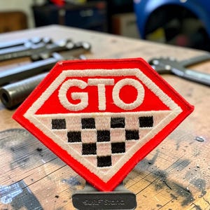May include: A diamond-shaped embroidered patch featuring a red background with the white text GTO centred at the top. The bottom half of the patch displays a black and white checkered pattern. The patch is mounted on a small black stand.