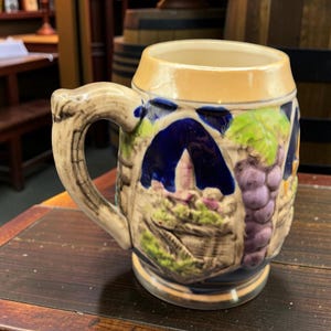 Peut inclure: Une chope à bière en céramique avec une anse, présentant un motif en relief détaillé d'une scène de vignoble avec des raisins, un bâtiment et du feuillage dans des tons de bleu, vert, violet et beige. La chope a un bord et une anse beige clair.