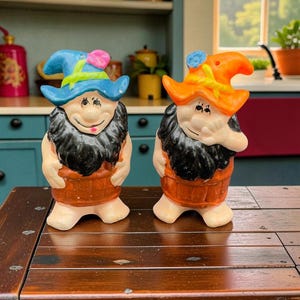 Peut inclure: Deux figurines de gnome en céramique avec des barbes noires, des barils orange et des chapeaux colorés. L'un porte un chapeau bleu avec une fleur rose, l'autre un chapeau orange. Ils sont sur une table en bois.