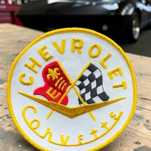 May include: A circular embroidered patch featuring the Chevrolet Corvette logo with yellow text, a red flag with a gold fleur-de-lis, and a black and white checkered flag on a white background, all enclosed within a thick yellow stitched border.