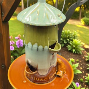 May include: A hanging ceramic bird feeder with a light green roof and a brown base. The feeder has a cylindrical body with openings for birds to access food. The feeder is suspended by a white cord.