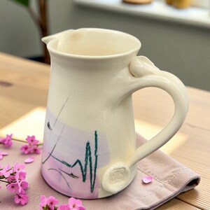 Peut inclure: Pichet en céramique crème avec une anse et un bec courbés. Le pichet présente un motif peint avec des accents violets et verts. Des fleurs et des pétales roses sont dispersés autour du pichet sur un tissu rose.