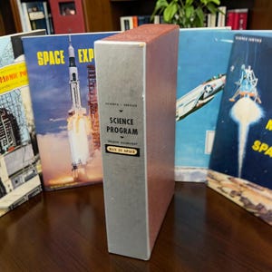 Peut inclure: Une collection de livres vintage Science Program sur le thème de l'exploration spatiale, incluant des titres comme Man in Space et Space Exploration, accompagnés d'un modèle de fusée rouge et blanc sur une surface en bois sombre.