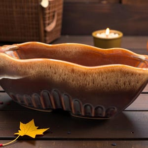 Peut inclure: Un plat de service en céramique marron et beige avec un bord ondulé et des rainures décoratives. Le plat a une finition brillante et est probablement utilisé pour servir des aliments. Le fond comprend des décorations sur le thème de l'automne.