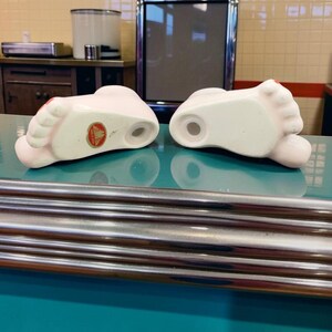 May include: Two ceramic foot-shaped salt and pepper shakers with a pale pink finish and red painted toenails. Each shaker features a small circular opening on the top and a round hole on the bottom, with one shaker displaying a circular gold sticker.