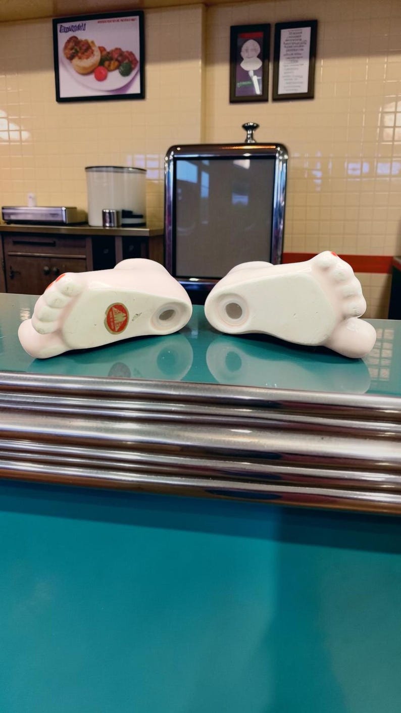 May include: Two ceramic foot-shaped salt and pepper shakers with a pale pink finish and red painted toenails. Each shaker features a small circular opening on the top and a round hole on the bottom, with one shaker displaying a circular gold sticker.