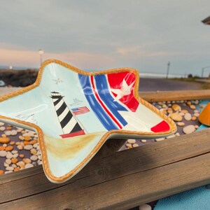 May include: A star-shaped ceramic bowl featuring a painted lighthouse, a small American flag, and red, white, and blue stripes. The bowl has a light blue background and a textured, rope-like trim along the outer edge of its five points.