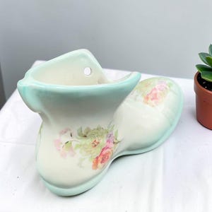 Peut inclure: Un pot de fleurs en céramique en forme de chaussure avec une base crème, un bord et un talon vert menthe pâle. L'extérieur est orné d'un motif floral composé de fleurs roses, orange et vertes. Un petit trou circulaire est visible près du bord.