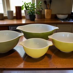 Peut inclure: Quatre saladiers Pyrex vintage, verts et jaunes, posés sur une table en bois. Les bols ont un design classique avec un intérieur blanc et une forme arrondie, parfaits pour la cuisine.