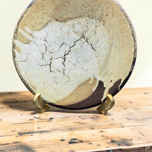 Peut inclure: Une assiette ronde en céramique présentant une surface beige et blanche tachetée avec un motif de glaçure craquelée. La partie inférieure de l'assiette affiche une couleur de base brun foncé avec un effet de coulure, sur un support en métal doré.