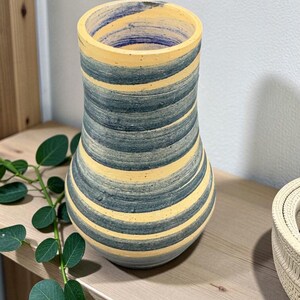 May include: A striped ceramic vase in yellow, blue, and grey. The vase has a wide opening and a rounded base. It is displayed on a wooden shelf with a trailing green plant. The vase is approximately 20 cm tall.