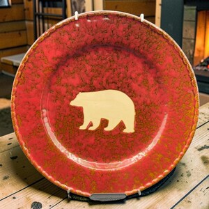 Peut inclure: Une grande assiette décorative rouge et ronde avec une silhouette d'ours crème au centre. L'assiette est présentée sur une table en bois. L'arrière-plan comprend une cheminée et des poteries.