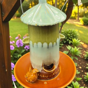 Peut inclure: Un nichoir en céramique aux tons de vert clair, marron et orange. Il est doté d'un toit en forme de dôme et d'une base circulaire avec une figurine de petit oiseau. Suspendu par un cordon blanc, il se détache sur un fond vert flou.