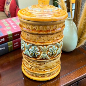 Peut inclure: Un pot en céramique avec un couvercle, présentant une palette de couleurs chaudes de jaune, marron et bleu. Le pot a une forme cylindrique avec des motifs floraux et tourbillonnants décoratifs. Le couvercle a un petit bouton. Le pot mesure environ 25 cm de haut.