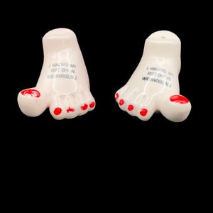 May include: A pair of ceramic salt and pepper shakers shaped like human feet with red painted toenails. Each foot features the black printed text I WALKED MY FEET OFF IN WILDWOOD, N.J. on the top surface, with small holes for dispensing seasoning.