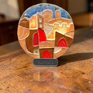 May include: A circular ceramic plaque featuring a colorful, stylized cityscape with a blue sky, tan buildings, and vibrant red accents. The piece is supported by a small, dark gray rectangular stand labeled with the text SURF Stand.