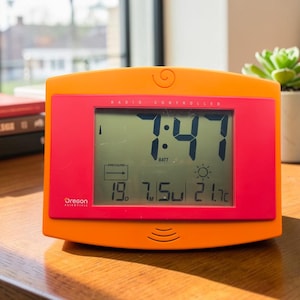 May include: An orange and pink digital alarm clock with a rectangular screen displaying the time 7:47, a sun icon, and temperature readings of 19.7 and 21.7 degrees Celsius. The device features the text Oregon Scientific on the lower left corner.