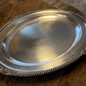 May include: Oval silver serving tray with a decorative rope-like border and floral accents. The tray sits on a wooden surface, with a glass of amber liquid in the background. A linen napkin is also visible.