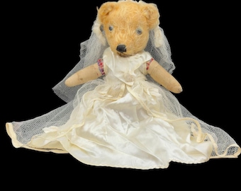 May include: A vintage teddy bear dressed in a white satin wedding dress and a delicate veil. The bear has light brown fur and is set against a black backdrop. The dress has a layered skirt and lace details, creating a classic bridal appearance.