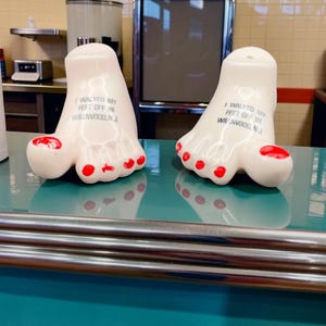 May include: Two white ceramic salt and pepper shakers shaped like human feet with red painted toenails. Each shaker features the black printed text I WALKED MY FEET OFF IN WILDWOOD, N.J. on the top surface of the foot-shaped ceramic body.