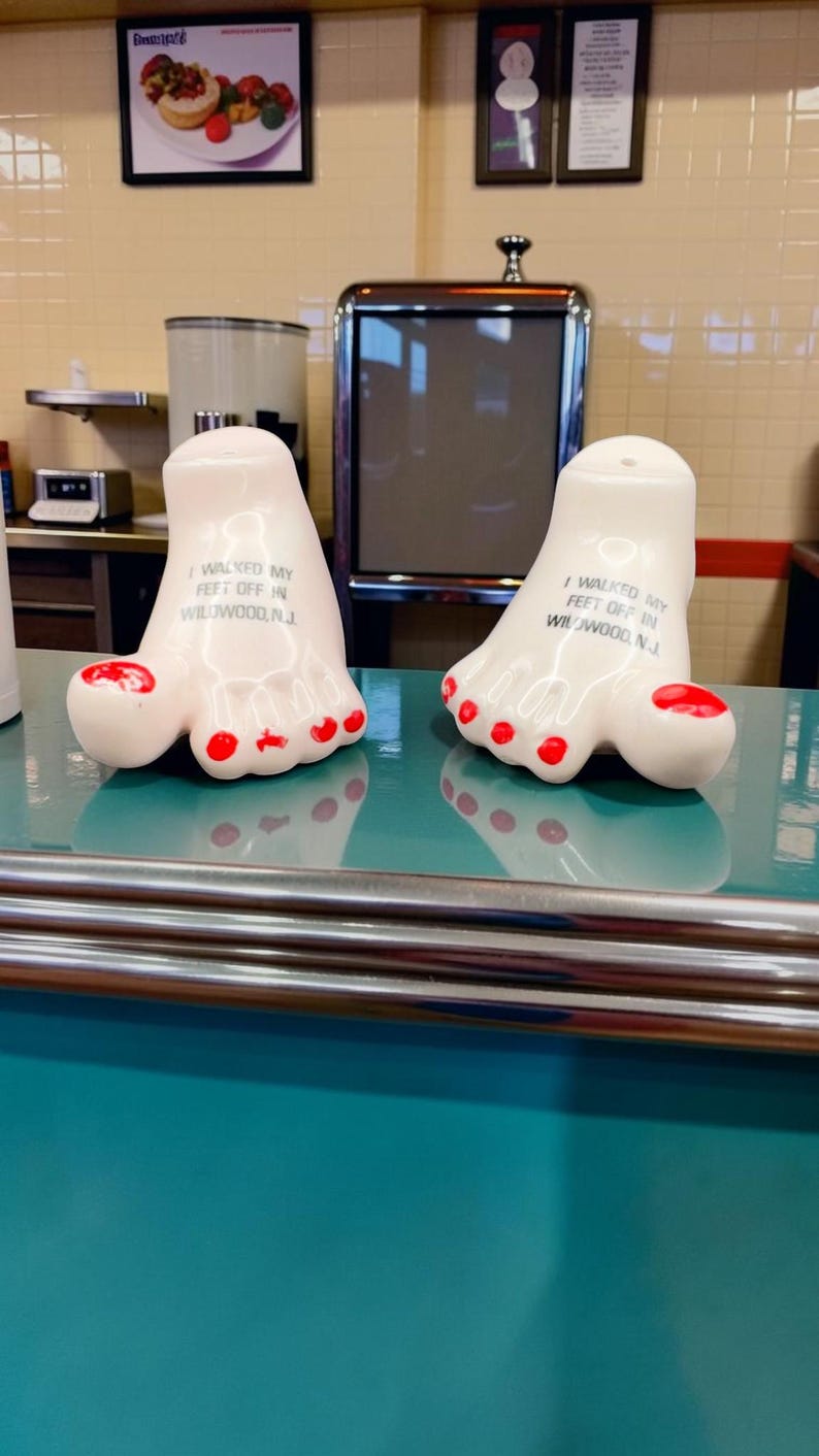 May include: Two white ceramic salt and pepper shakers shaped like human feet with red painted toenails. Each shaker features the black printed text I WALKED MY FEET OFF IN WILDWOOD, N.J. on the top surface of the foot-shaped ceramic body.