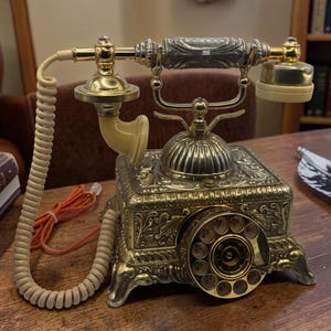 May include: A detailed antique brass telephone with a cream-coloured handset and cord. The rotary dial phone is on a wooden desk. A vintage book and a quill pen are also on the desk, suggesting a classic office setting. The phone is a decorative item.