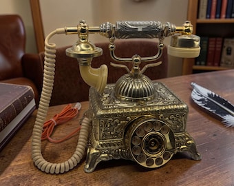 May include: A detailed antique brass telephone with a cream-coloured handset and cord. The rotary dial phone is on a wooden desk. A vintage book and a quill pen are also on the desk, suggesting a classic office setting. The phone is a decorative item.