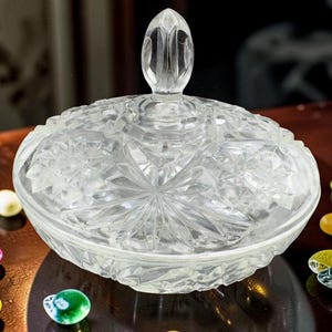 Peut inclure: Un bonbonnière en verre taillé transparent avec un couvercle, présentant un motif en étoile sur la base et le couvercle. Le plat est de forme ovale et repose sur une surface sombre, entouré de bonbons colorés de forme ovale.