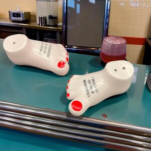 May include: Two white ceramic foot-shaped salt and pepper shakers with red painted toenails. Each shaker features the black printed text I WALKED MY FEET OFF IN WILDWOOD, N.J. The shakers have small holes on the top for dispensing seasoning.