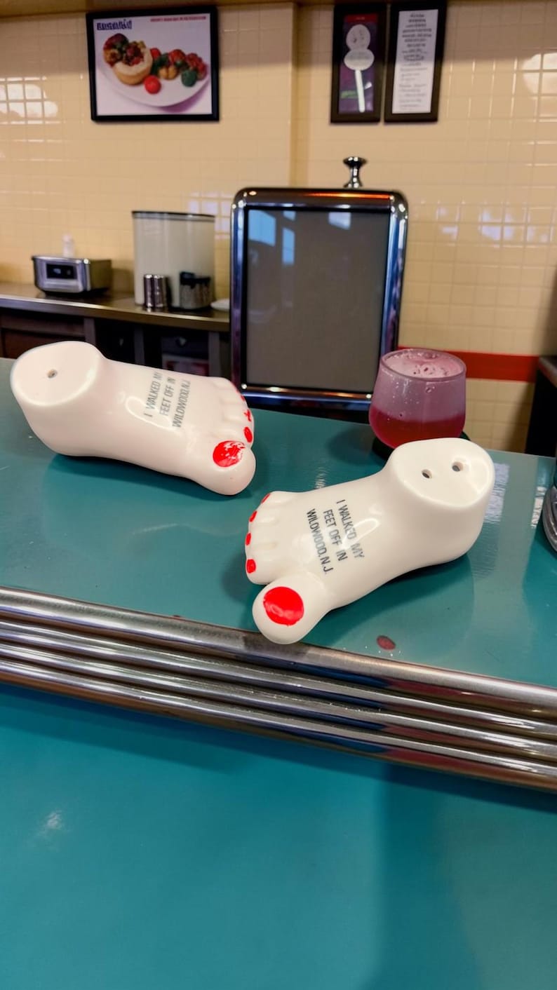 May include: Two white ceramic foot-shaped salt and pepper shakers with red painted toenails. Each shaker features the black printed text I WALKED MY FEET OFF IN WILDWOOD, N.J. The shakers have small holes on the top for dispensing seasoning.