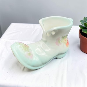 May include: A pale mint green ceramic shoe-shaped planter with a glossy finish. The piece features delicate hand-painted floral motifs in soft pink and yellow hues on the toe and side, with a smooth, curved silhouette and a wide opening at the top.