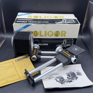 May include: A Soligor Flexomatic double track bellows unit for macro photography, featuring black accordion-style bellows and silver metal rails. The set includes the original white and blue cardboard box, a yellow instruction sheet, and a white manual.