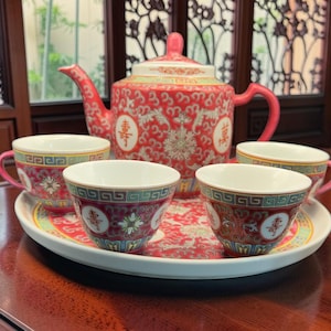 May include: A ceramic tea set featuring a teapot and four matching cups arranged on a round tray. The pieces are decorated with a vibrant red pattern, intricate gold-toned floral motifs, and Chinese characters, finished with a teal geometric border.
