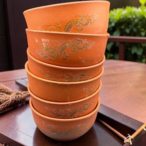 Peut inclure: Une pile de six bols de couleur terre cuite avec un motif floral doré délavé. Les bols sont empilés sur un plateau en bois foncé, suggérant un ensemble pour servir ou exposer. Les bols ont un aspect rustique et fait à la main.