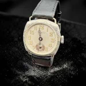 May include: Vintage wristwatch with a cream dial, black leather strap, and silver case. The watch has Roman numerals and a small seconds dial. The watch is set against a dark background.