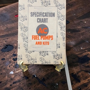 Peut inclure: Un tableau de spécifications vintage en papier beige pour les pompes à carburant et kits AC. La couverture présente un motif répétitif de dessins au trait bleu foncé de pompes à carburant, avec un texte noir et orange indiquant Specification Chart.