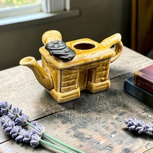 Peut inclure: Une théière fantaisiste de couleur beige, avec un design unique en forme de maison, avec une cheminée et une poignée. Deux objets noirs et circulaires sont posés sur le dessus. Sur une table en bois rustique, avec des brins de lavande et des livres anciens.