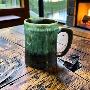 Peut inclure: Une tasse en céramique dotée d'un corps cylindrique et d'une anse. L'extérieur présente un émail bicolore, passant d'une base noir profond à une finition vert émeraude vif et dégoulinant sur le bord, créant un aspect texturé et superposé.