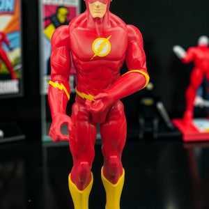 May include: A red action figure of The Flash with yellow lightning bolt emblems on the chest, forearms, and waist. The figure features a full-body red suit, yellow boots, and a red cowl with yellow ear wings, standing in a neutral pose.