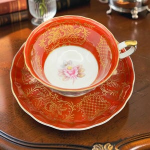 May include: A porcelain teacup and saucer set featuring a vibrant red glaze with intricate gold filigree patterns. The interior of the cup and the centre of the saucer display a delicate pink rose motif. The cup has a white handle with gold trim.