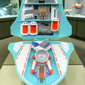 Peut inclure: Un ensemble de jeu vaisseau spatial en plastique bleu clair qui s'ouvre pour révéler un intérieur détaillé. La section supérieure présente un cockpit avec des panneaux de contrôle et une petite figurine. La section inférieure inclut un radar argenté.