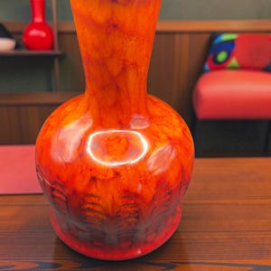 Peut inclure: Un vase en céramique bulbeux présentant un glaçage marbré orange et rouge vif. Le col est fin et s'évase légèrement au niveau de l'ouverture. La partie inférieure du corps affiche un motif en relief répétitif de lignes verticales et de creux.