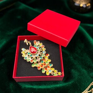 Peut inclure: Une broche paon dorée ornée d'une grande pierre centrale rouge en forme de goutte, entourée de petits cristaux multicolores dans des tons bleu, vert, jaune et orange, présentée dans une boîte cadeau carrée rouge ouverte avec un insert noir.